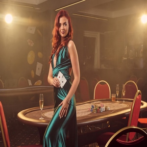 Alluring redheaded girl in a long blue satin dress is smiling and posing sideways with two aces in her hand against a poker table in luxury casino. Passion, cards, chips, alcohol, win, gambling - it is as female entertainment. Smoke background.
